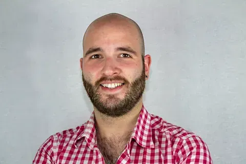 Un hombre calvo con barba viste una camisa a cuadros rojos y blancos y sonríe.