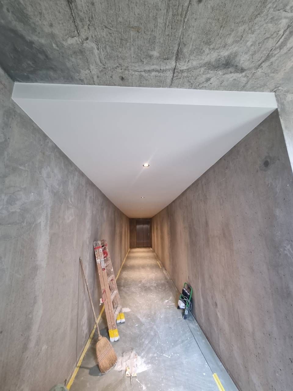 Couloir étroit en béton brut, avec un plafond blanc, outils et matériaux de construction le long du mur de gauche.