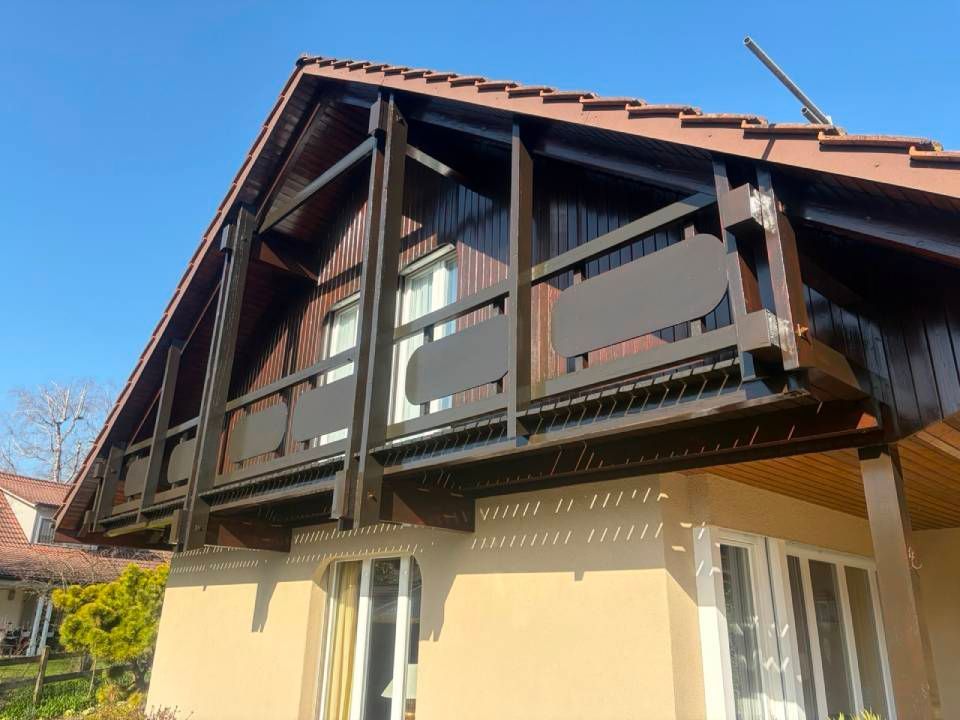 Maison à deux étages avec un balcon en bois sombre et des murs inférieurs beiges sous un ciel bleu clair
