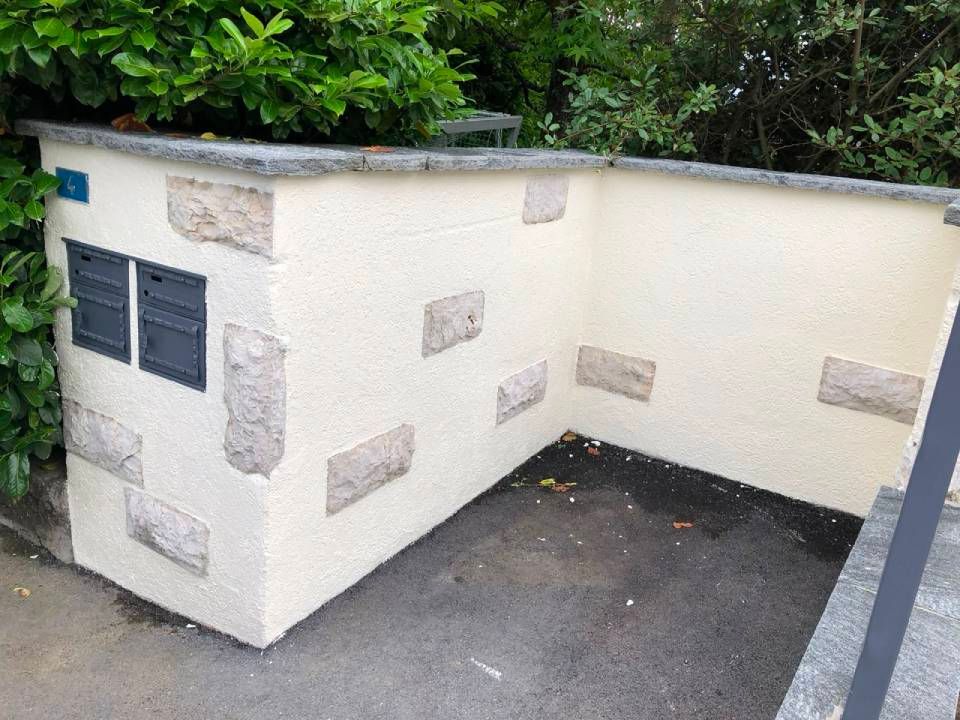 Bâtiment utilitaire d'angle blanc avec aérations sombres et accents de pierre, à côté d'un chemin pavé extérieur.