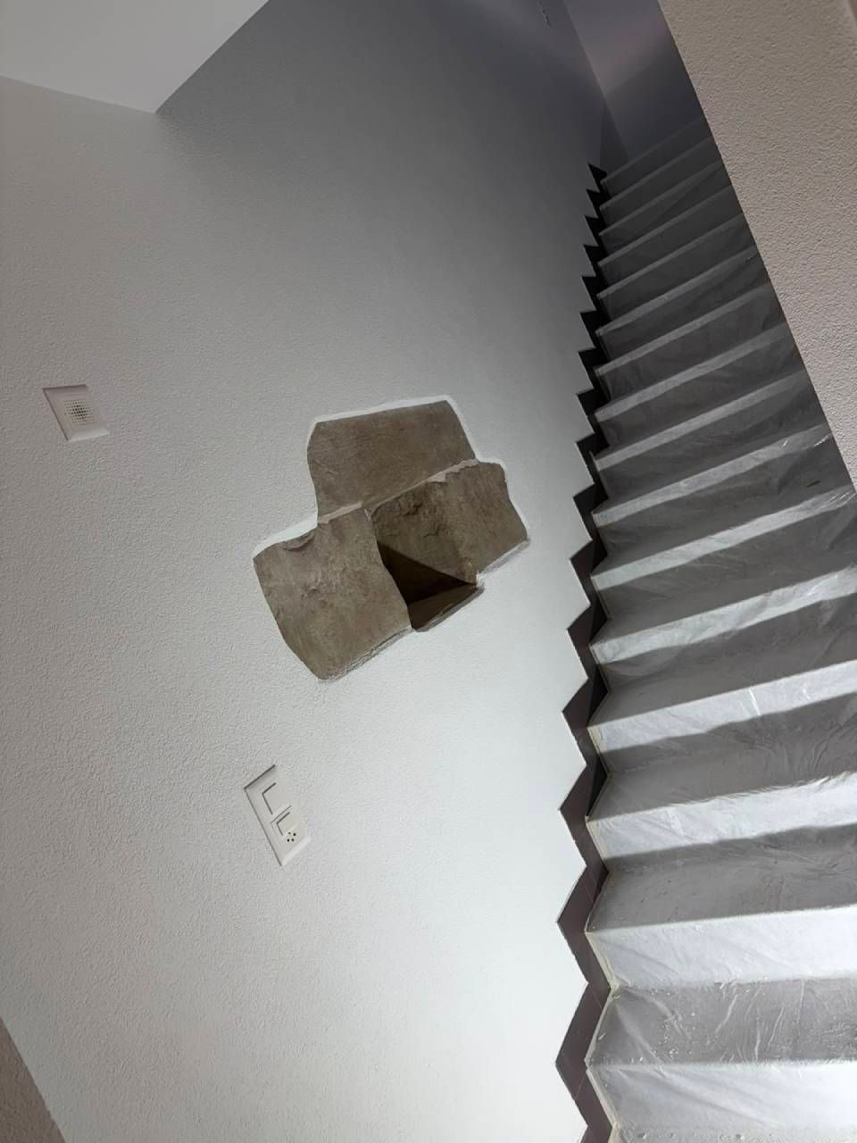 Mur intérieur incliné avec une petite ouverture carrée à côté d'un escalier.