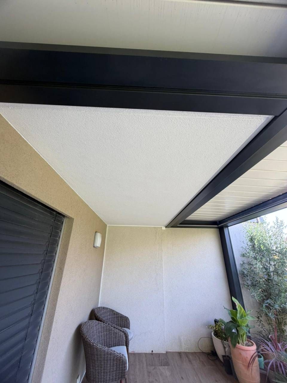 Patio couvert avec fauteuil en osier, plantes en pot et poutres apparentes noires au plafond, à côté d'une porte coulissante.