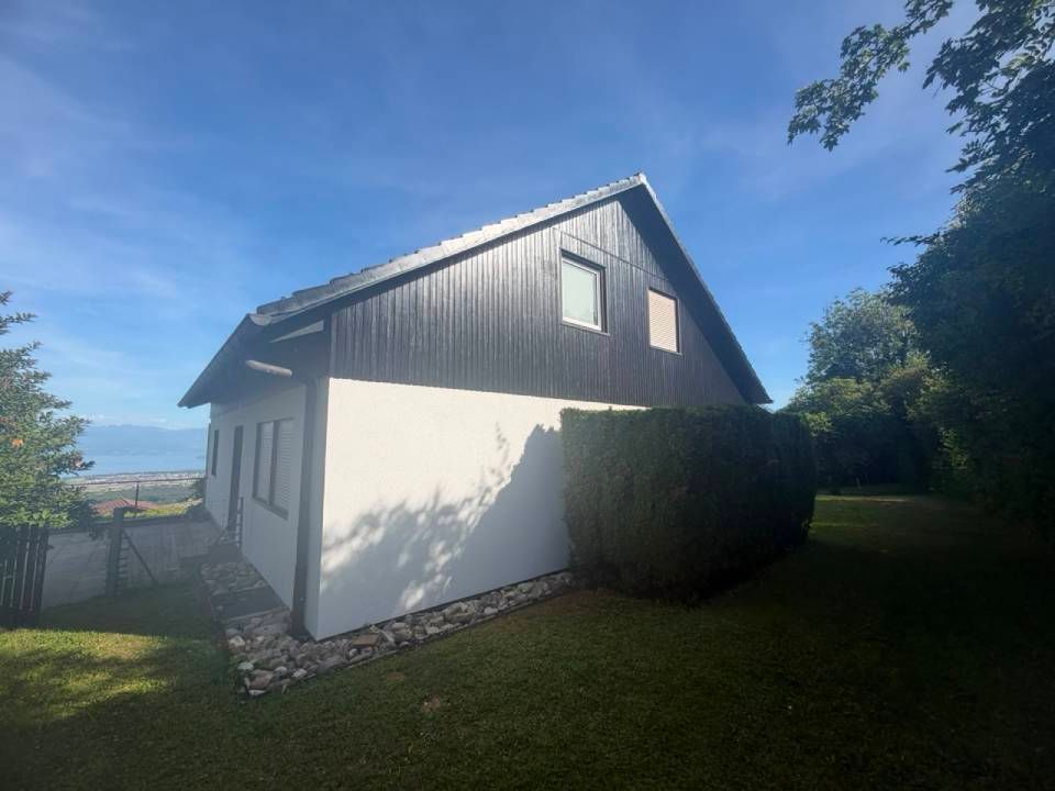 Maison blanche au toit sombre et à la haie, jouxtant une cour herbeuse sous un ciel bleu.