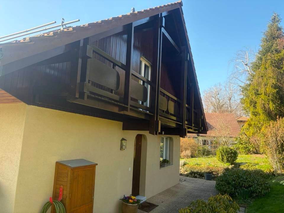 Maison couleur crème avec poutres de toit en bois foncé, jardinières et jardin baigné de soleil.