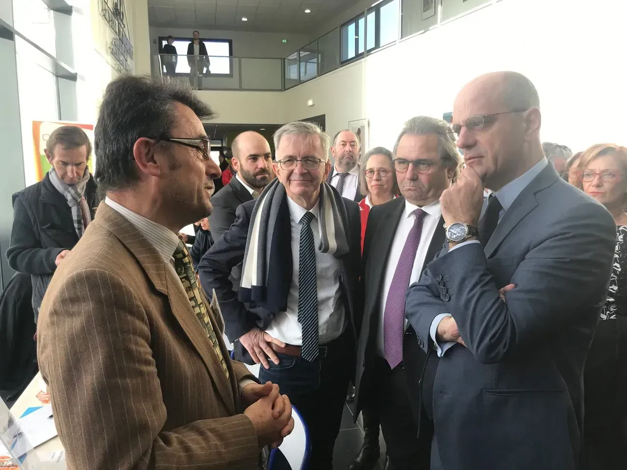 Des hommes en costume discutent dans un espace intérieur lumineux, probablement un hall d'entrée.