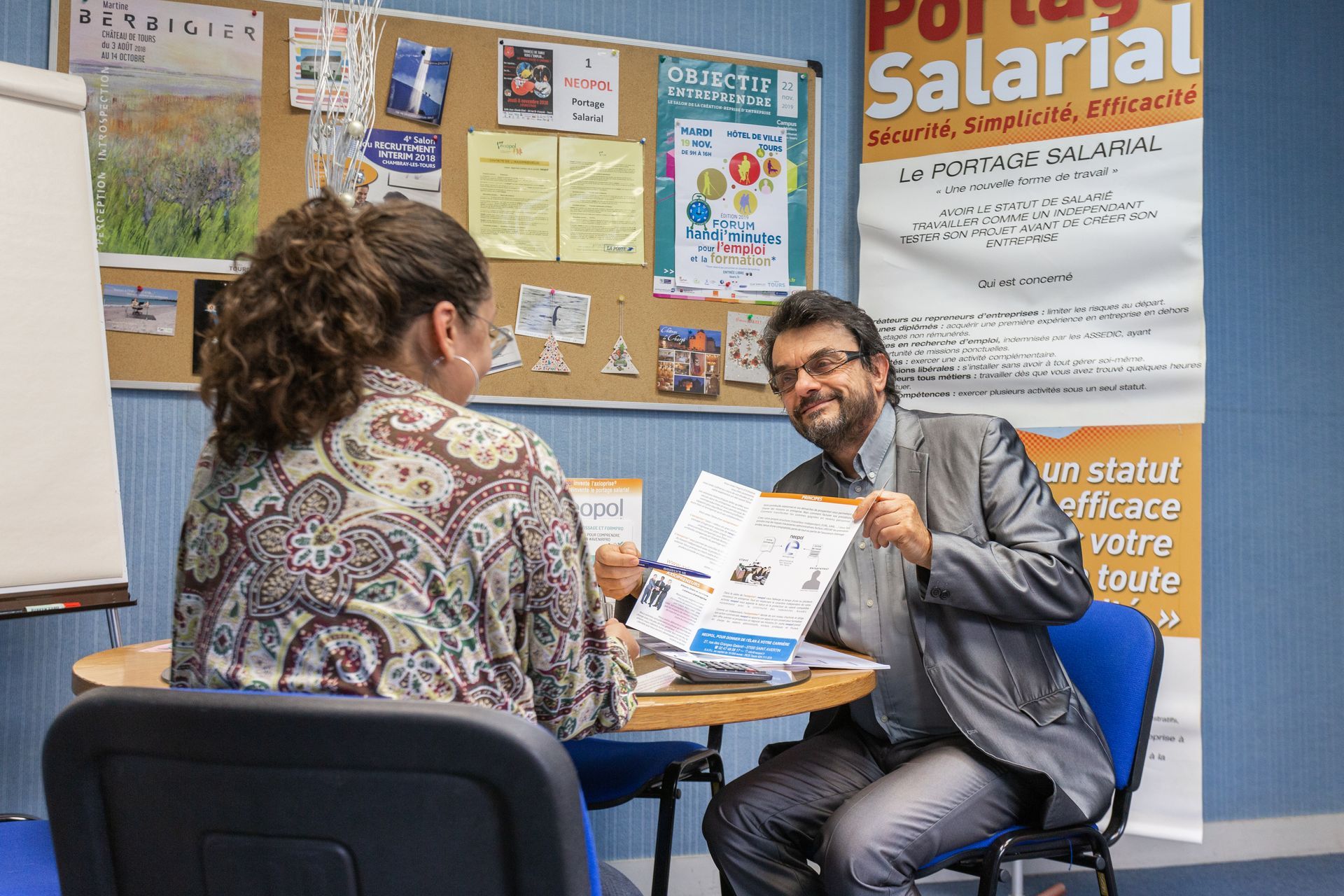 Un homme montre un document à une femme assise à une table. Ils se trouvent dans un bureau.