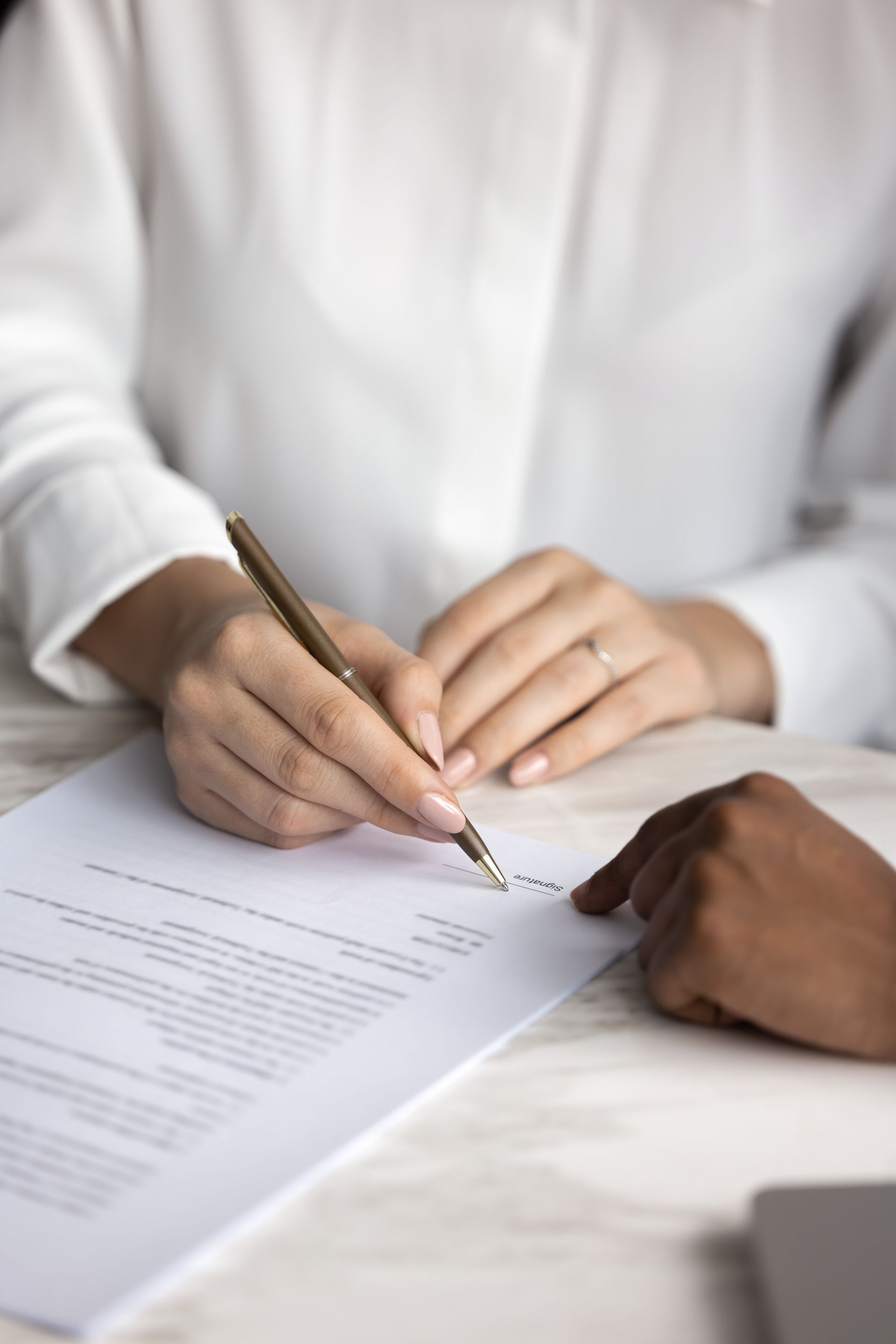 Une personne signe un document, une autre désigne du doigt. Chemise blanche, stylo, papier.