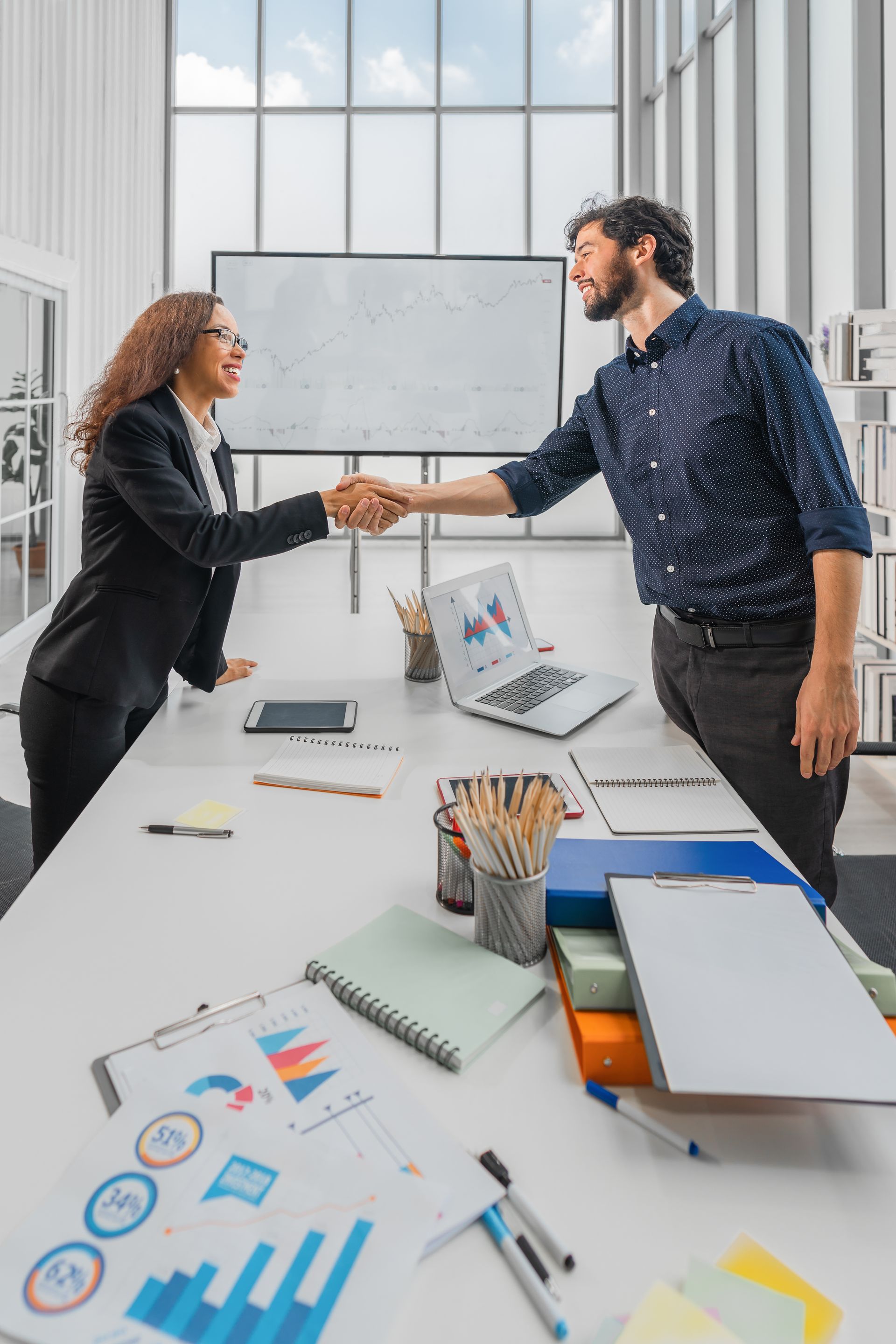 Un homme et une femme se serrent la main au-dessus d'un bureau, dans un environnement de bureau, avec un ordinateur portable et des documents.