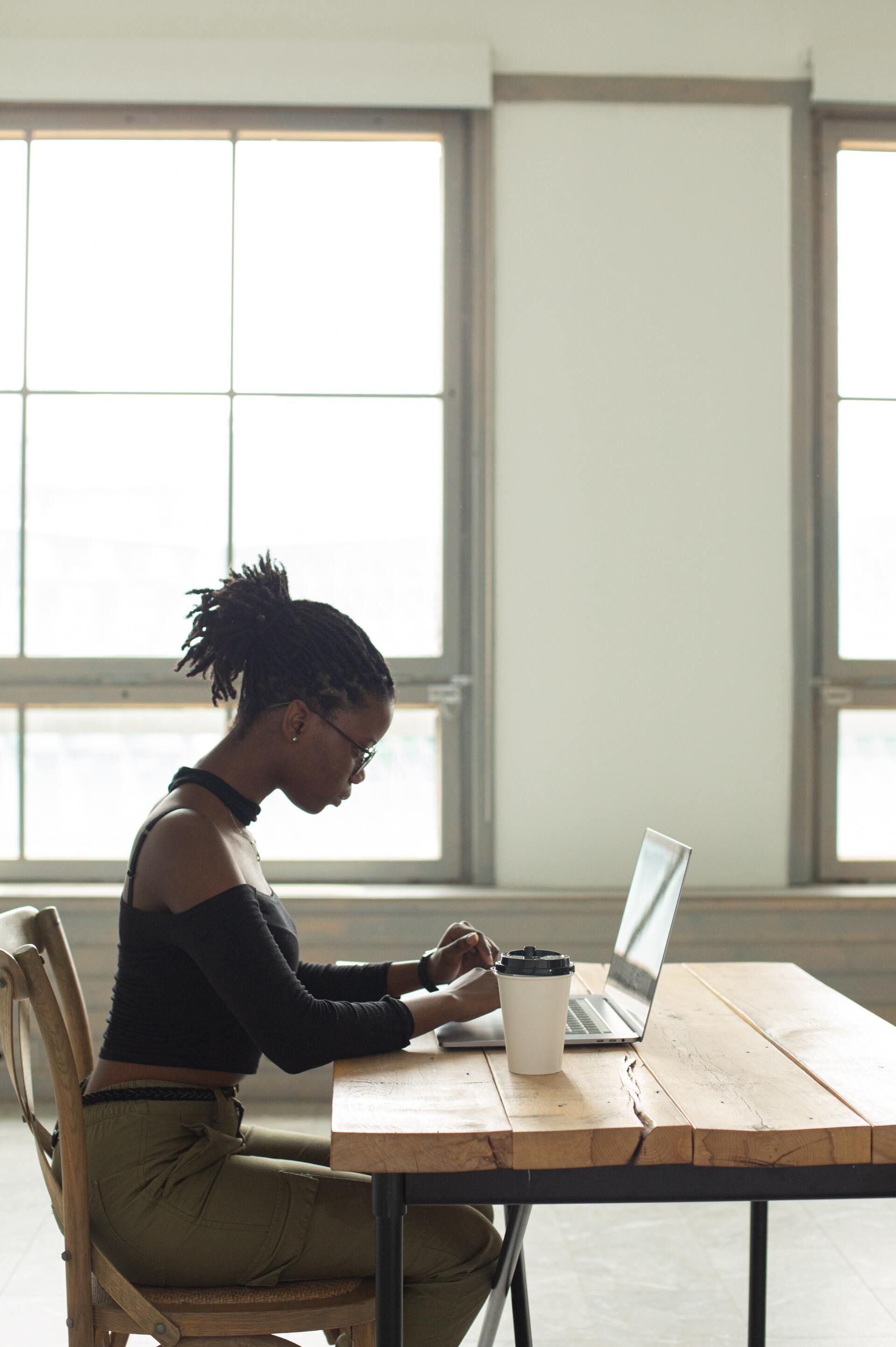 Une femme utilise un ordinateur portable à un bureau en bois près d'une fenêtre. Elle porte des lunettes et un haut noir.