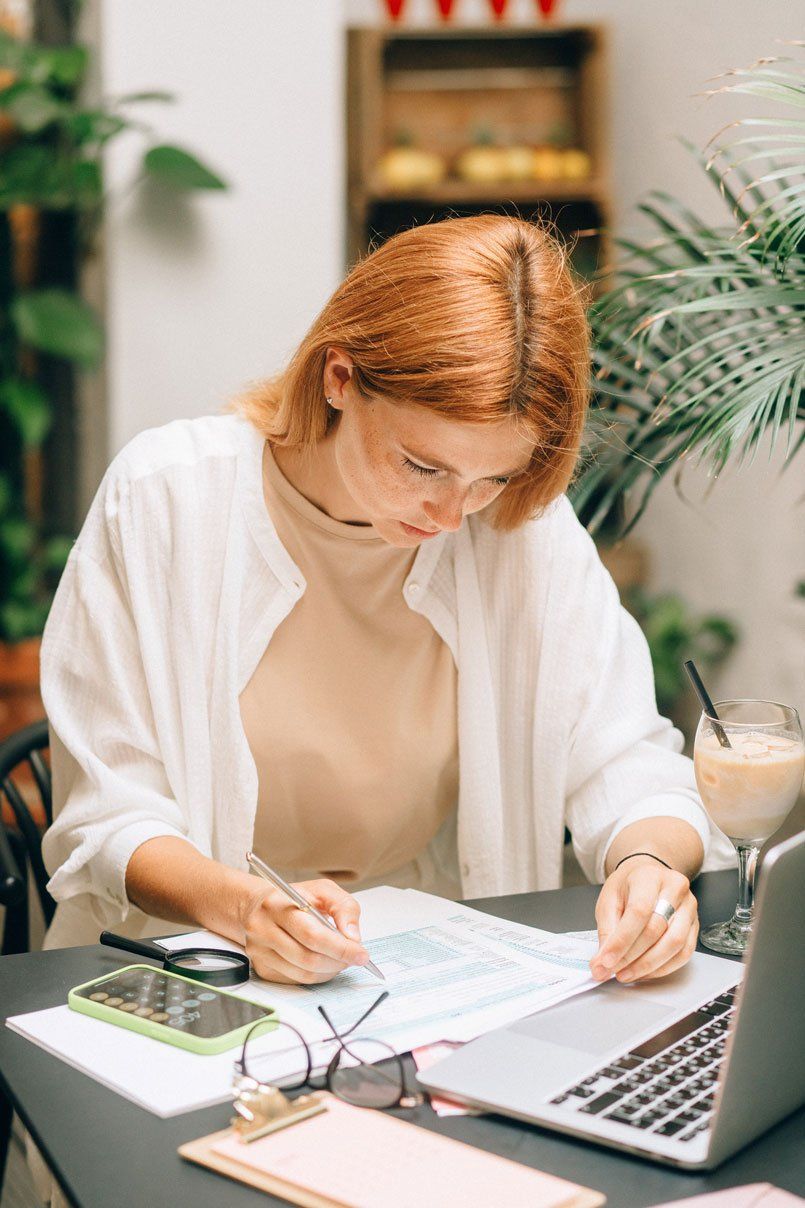 Une femme rousse, portant des lunettes, travaille sur des documents à une table avec un ordinateur portable, un téléphone et une boisson.