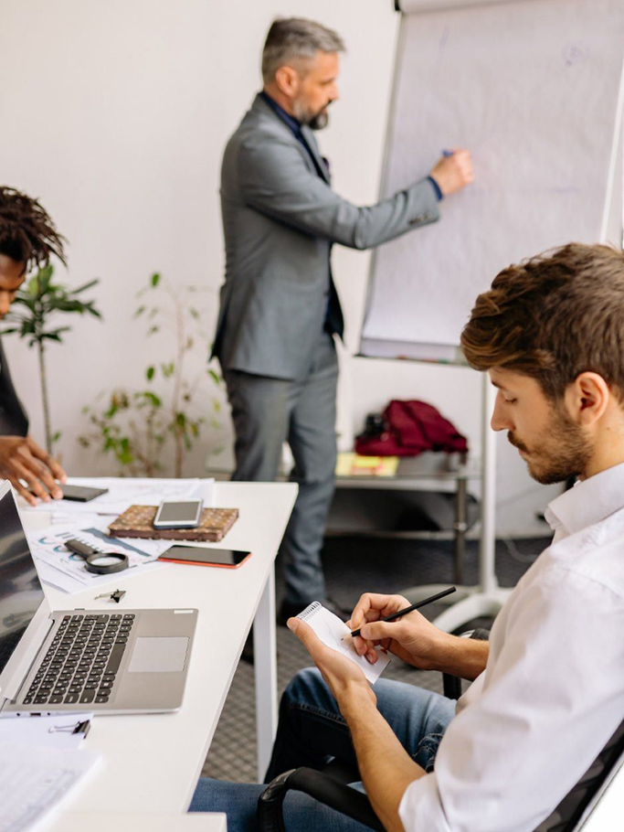 Un homme prend des notes pendant une réunion d'affaires, tandis qu'un collègue présente un exposé au tableau blanc. Deux autres personnes sont présentes.