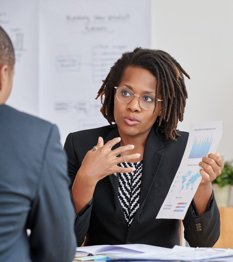 Une femme en blazer fait un geste, tenant un graphique. Elle est assise en face d'une personne en costume, dans un bureau.