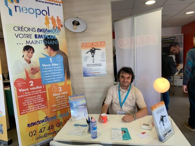 Un homme est assis à un stand portant la marque Neopal. Une banderole derrière lui fait la promotion de la création d'emplois.