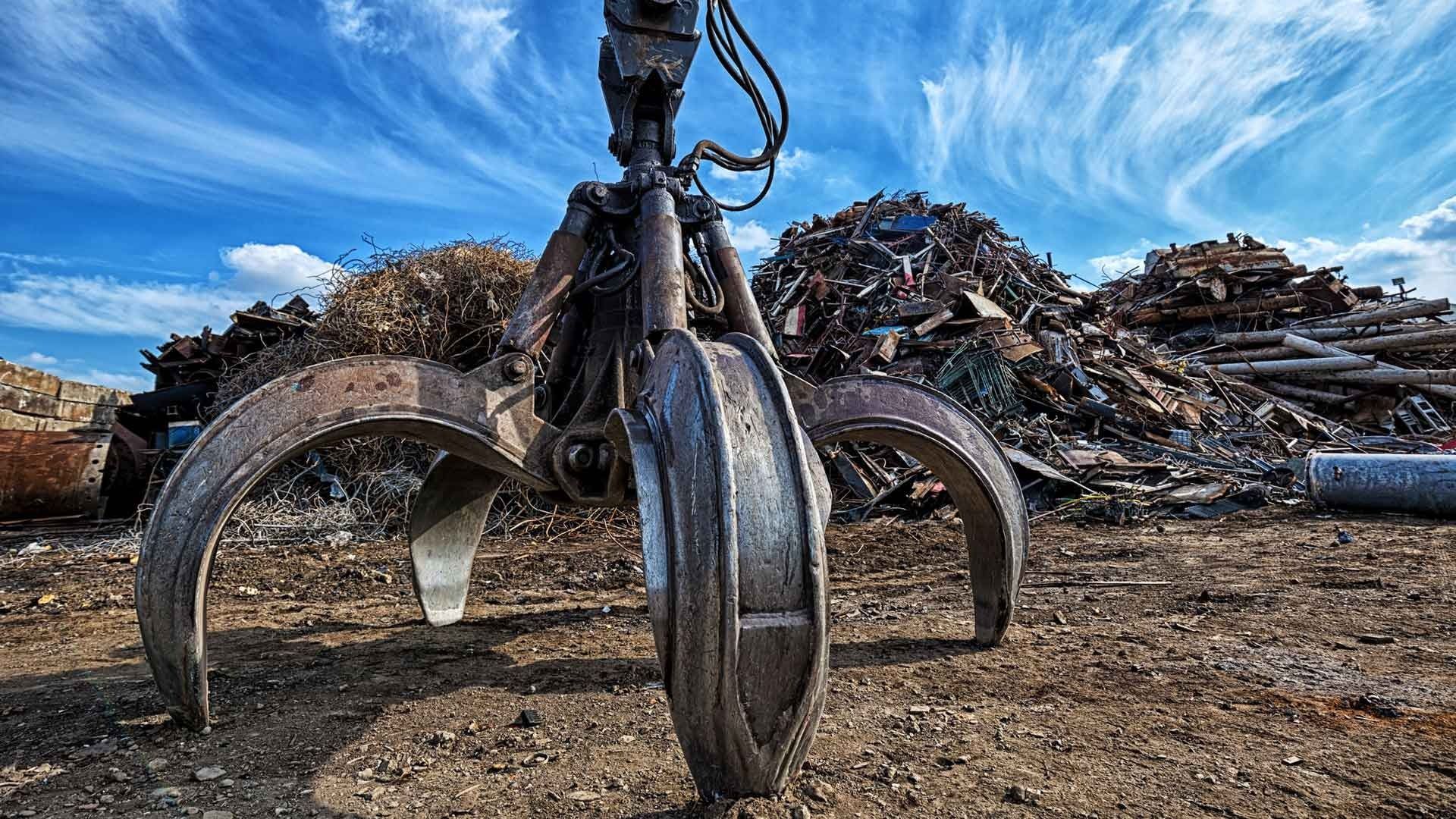 Gran garra de metal en un depósito de chatarra, sosteniendo una pila de metal bajo un cielo azul con nubes.