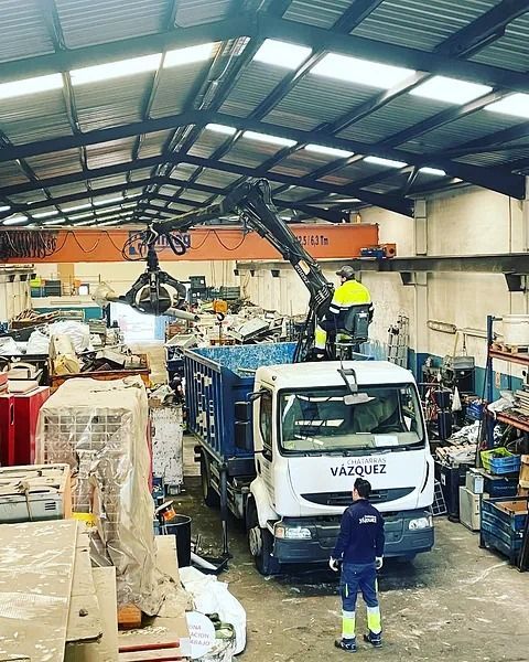Una grúa carga chatarra en un camión dentro de un almacén. Trabajadores con chalecos reflectantes.