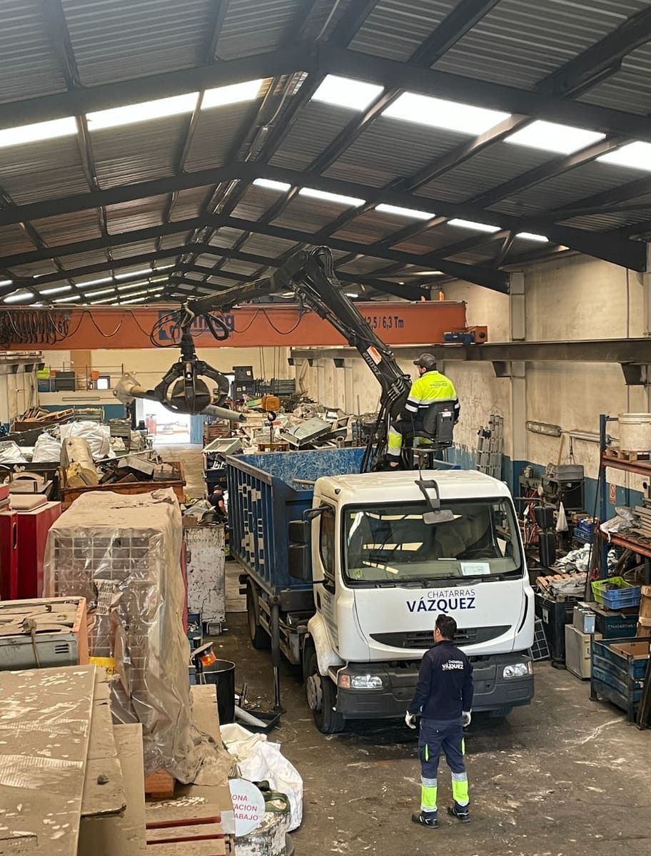 Un trabajador carga chatarra en un camión dentro de un almacén con una grúa.