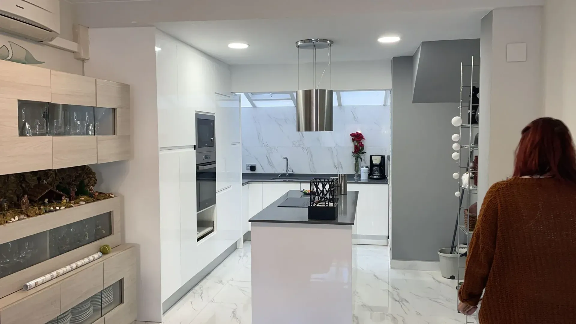 Cocina blanca moderna; la persona entra por la derecha. Isla, campana extractora de acero inoxidable, gabinetes con frente de vidrio y detalles en blanco y gris.