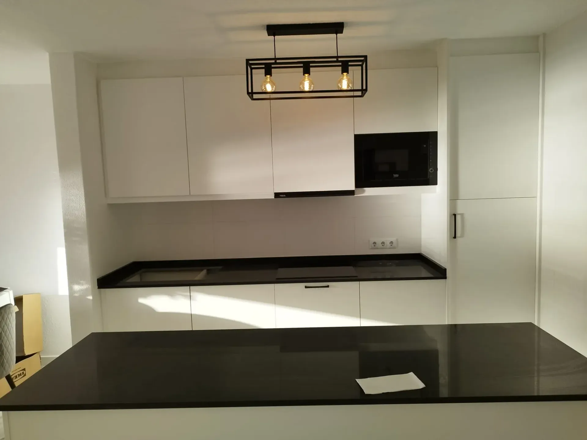 Cocina blanca con encimeras y gabinetes negros, y una lámpara rectangular. El microondas y la campana extractora son visibles.