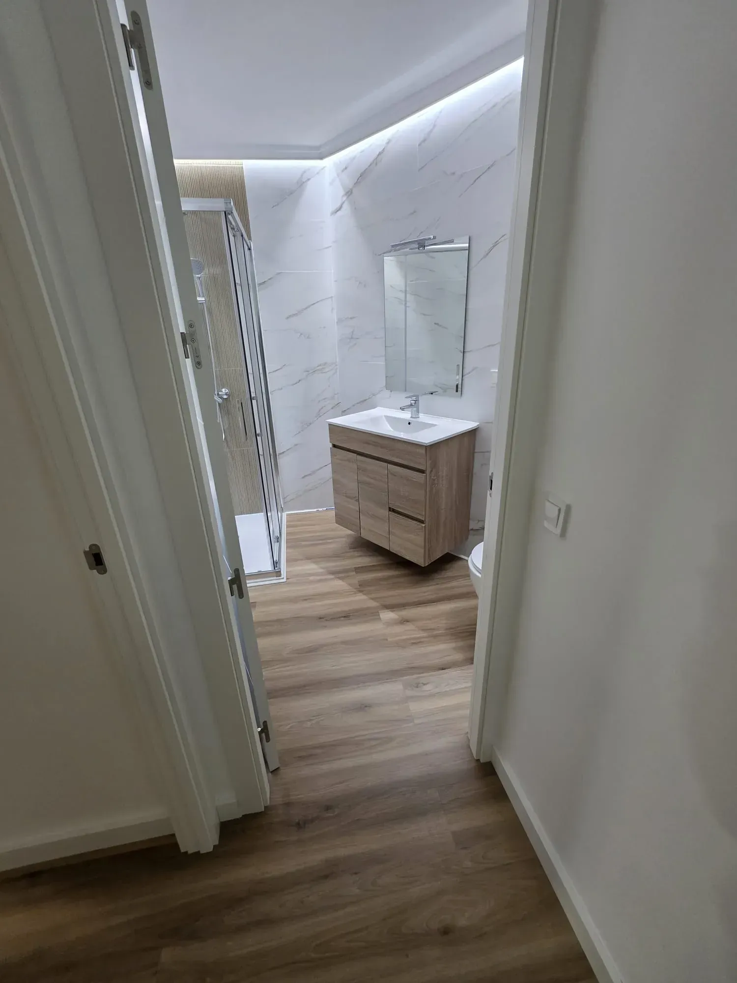 Interior de baño con piso de apariencia de madera, tocador, espejo y ducha, visto desde la puerta.