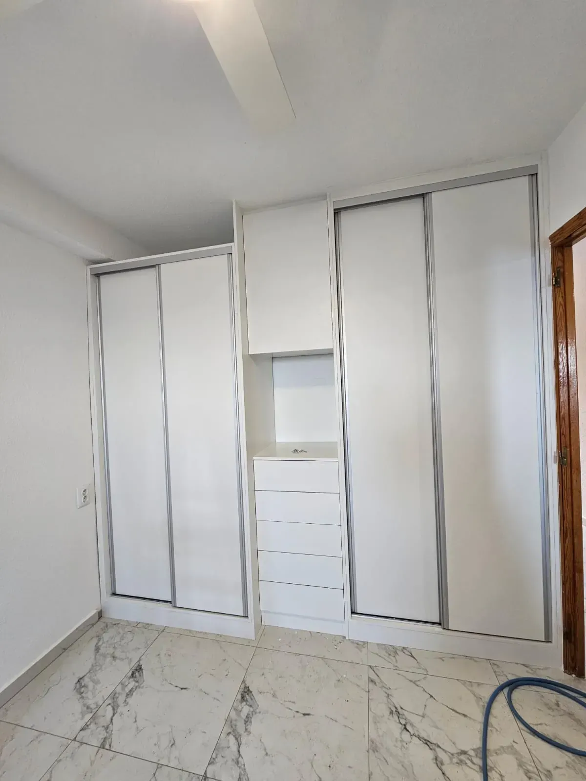 Armario blanco con puertas corredizas, detalles plateados y cajones empotrados contra una pared blanca en una habitación con piso de mármol.