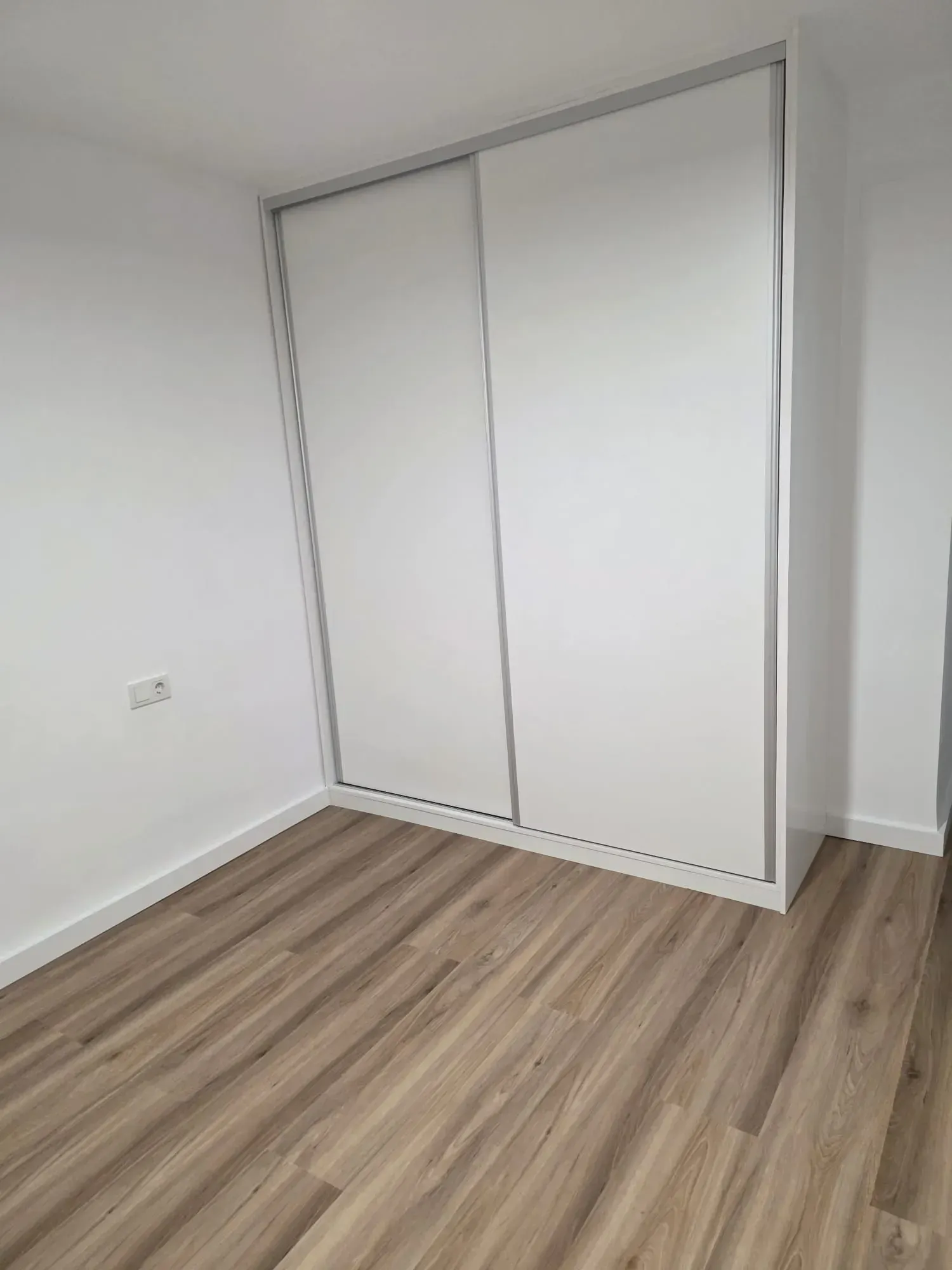 Armario blanco con puertas correderas en una habitación con suelo imitación madera clara y paredes blancas.