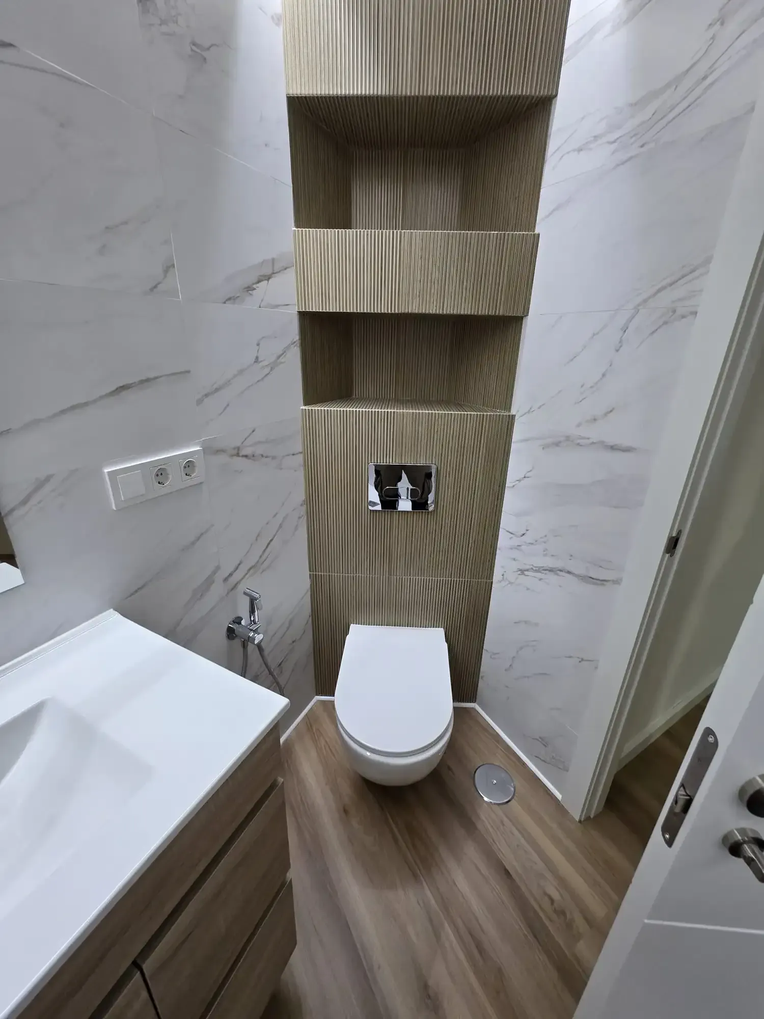 Baño moderno con texturas de madera y mármol: inodoro, lavabo y estantería empotrada.