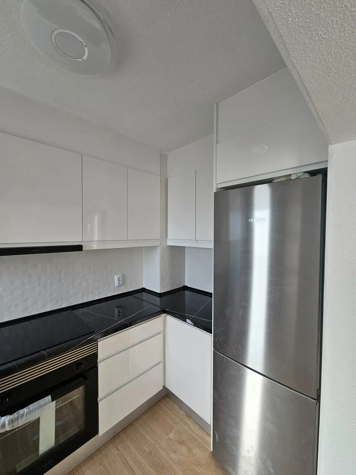 Pequeña cocina blanca con encimeras negras, refrigerador de acero inoxidable y horno.