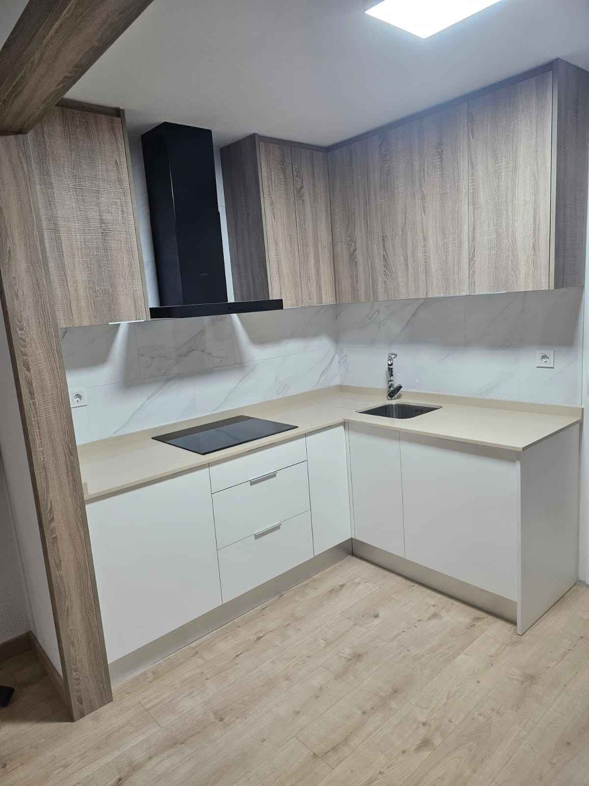 Cocina en forma de L con gabinetes de madera clara y blancos, campana extractora negra y encimeras claras.