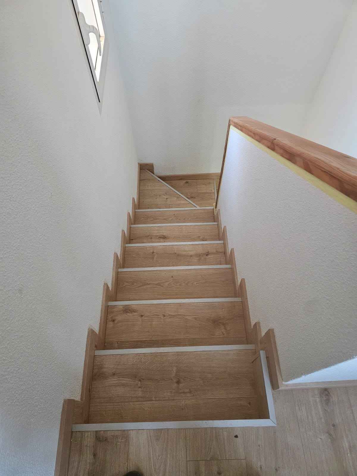 Escaleras de madera que suben, con pasamanos de madera a la derecha y paredes blancas.