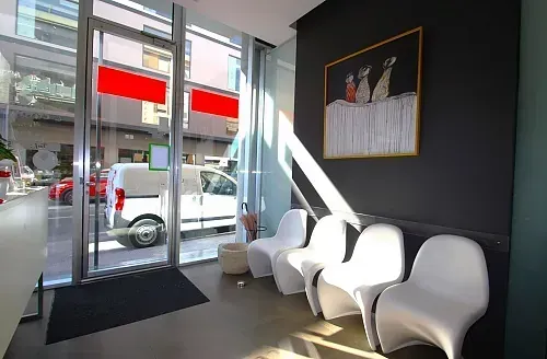 Sala de espera con sillas blancas, arte en una pared oscura y una puerta de vidrio que da a la calle.