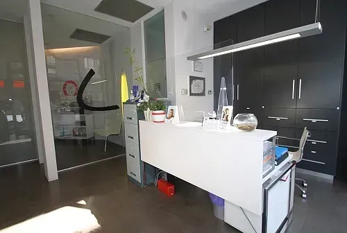 Área de recepción con un escritorio blanco, gabinetes negros y una puerta de vidrio a un área de espera.