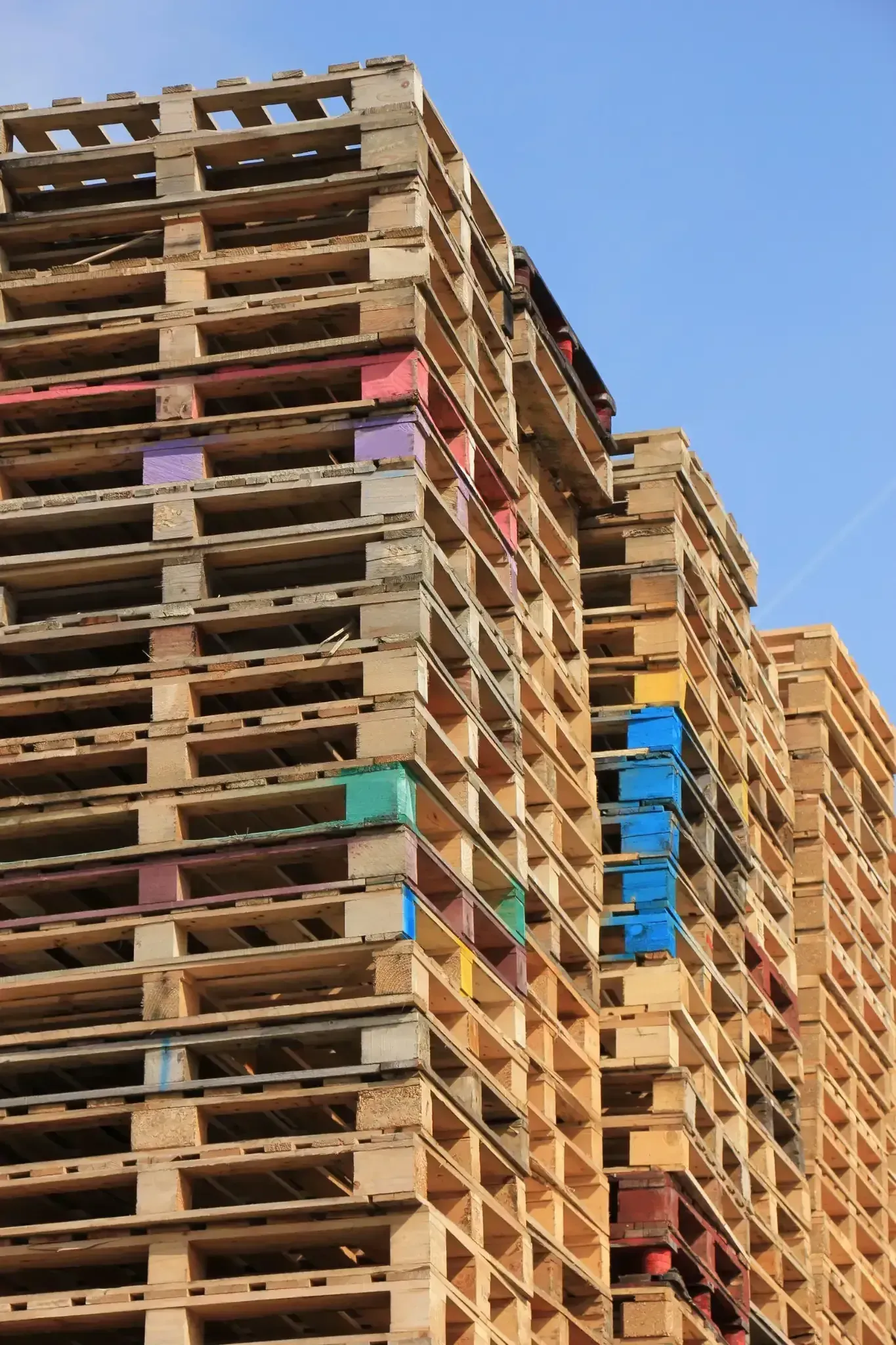 Paletas de madera apiladas, algunas con bloques de colores, contra un cielo azul.
