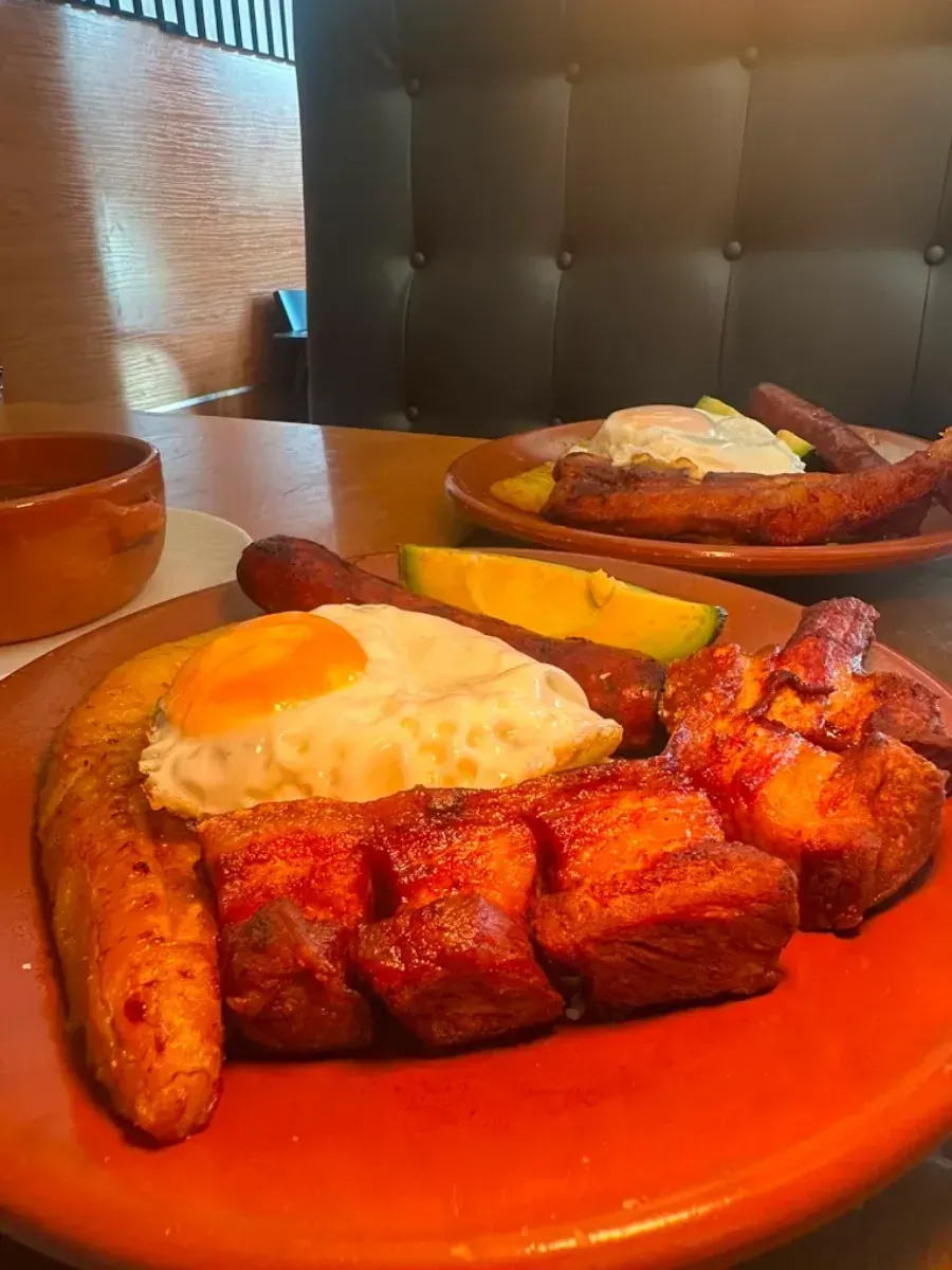 Plato de desayuno con huevos, salchicha, tocino, papas y tostadas en un plato naranja en una cabina de restaurante.