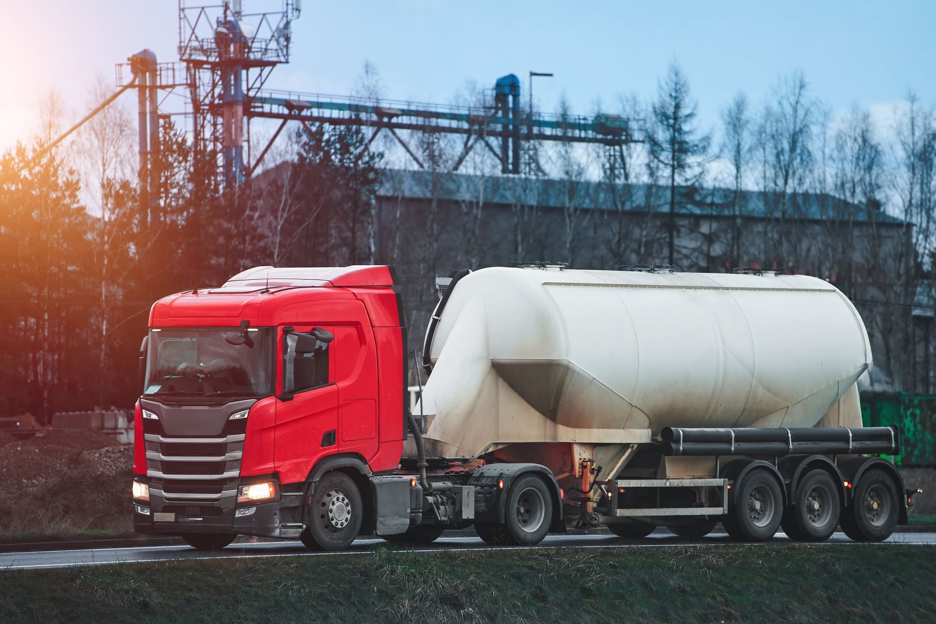 Un semi-remorque rouge avec une grande citerne blanche passe devant une zone industrielle au coucher du soleil.