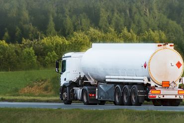 Un camion-citerne blanc circule sur une autoroute longeant une zone verdoyante bordée d'arbres.