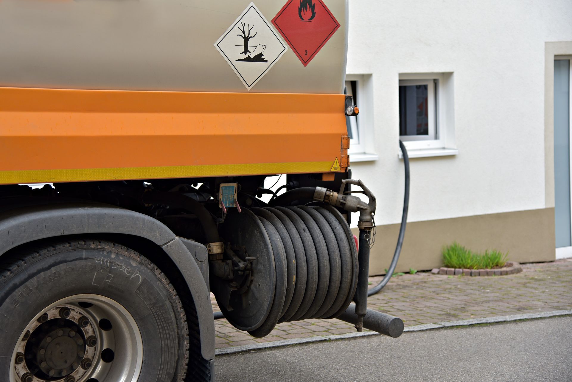 Un camion-citerne est stationné à côté d'un bâtiment, avec un enrouleur de tuyau visible et des panneaux de signalisation de danger sur le côté.