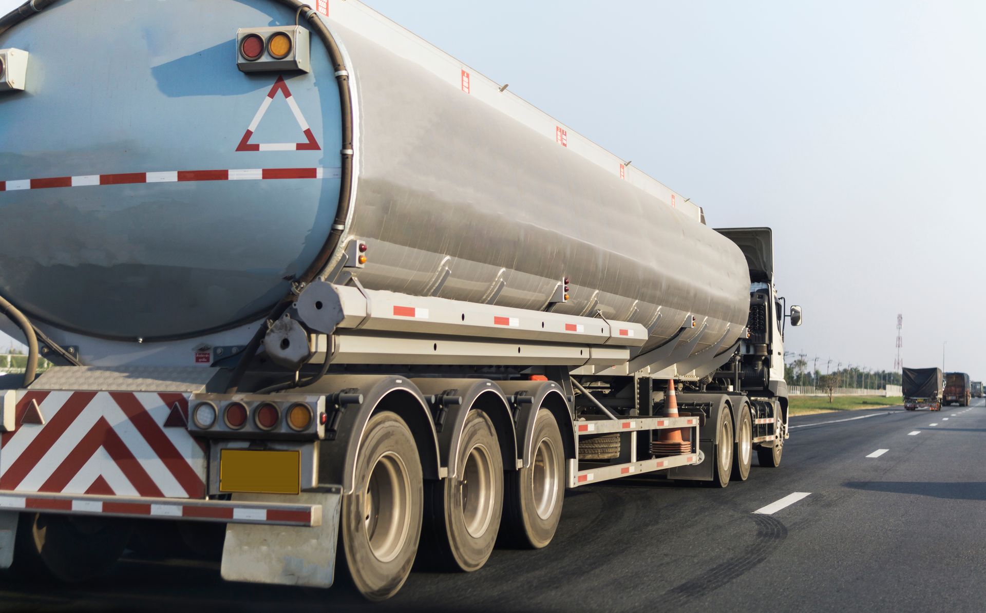 Un camion-citerne argenté circule sur une autoroute, vu de l'arrière avec des marquages de danger rouge et blanc.