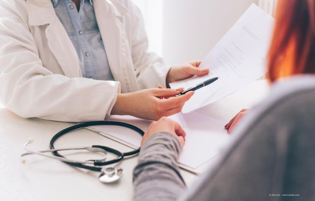 Der Arzt zeigt auf ein Dokument für den Patienten an einem Schreibtisch; das Stethoskop ist sichtbar.