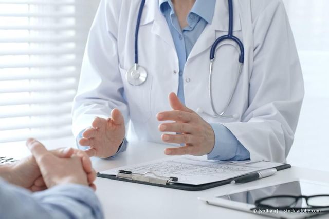 Arzt im weißen Kittel gestikuliert in Richtung Patient; Klemmbrett auf dem Schreibtisch, Stethoskop sichtbar.