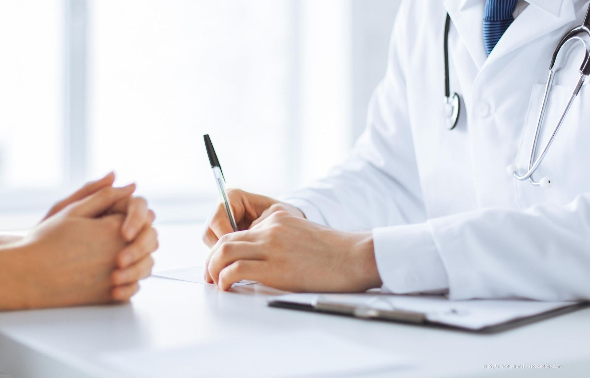 Der Arzt schreibt an einem Schreibtisch, die Hände des Patienten sind ihm gegenüber gefaltet.
