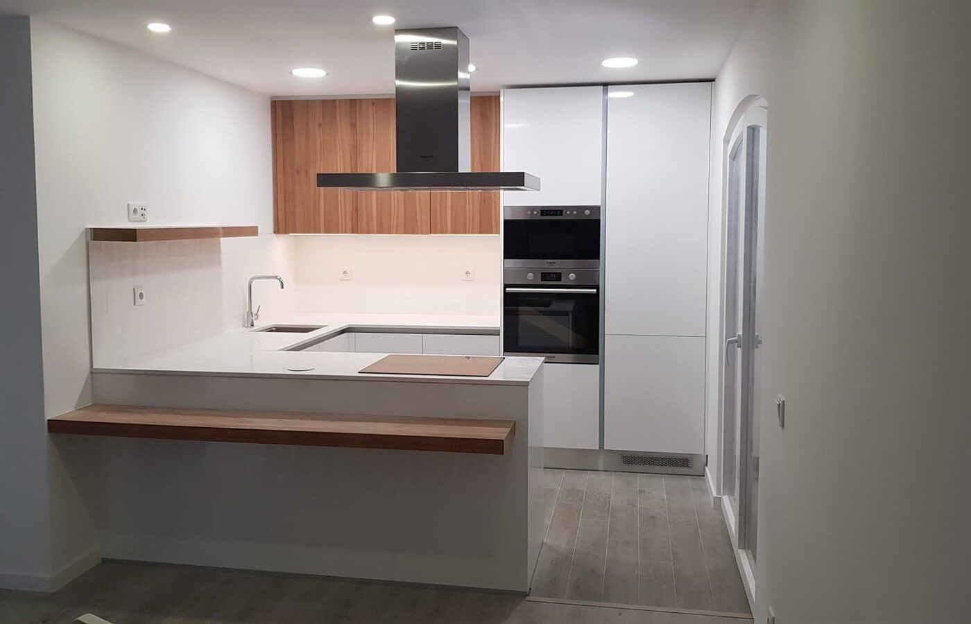 Cozinha moderna com armários brancos, detalhes em madeira, coifa de aço inoxidável e balcão para refeições rápidas.