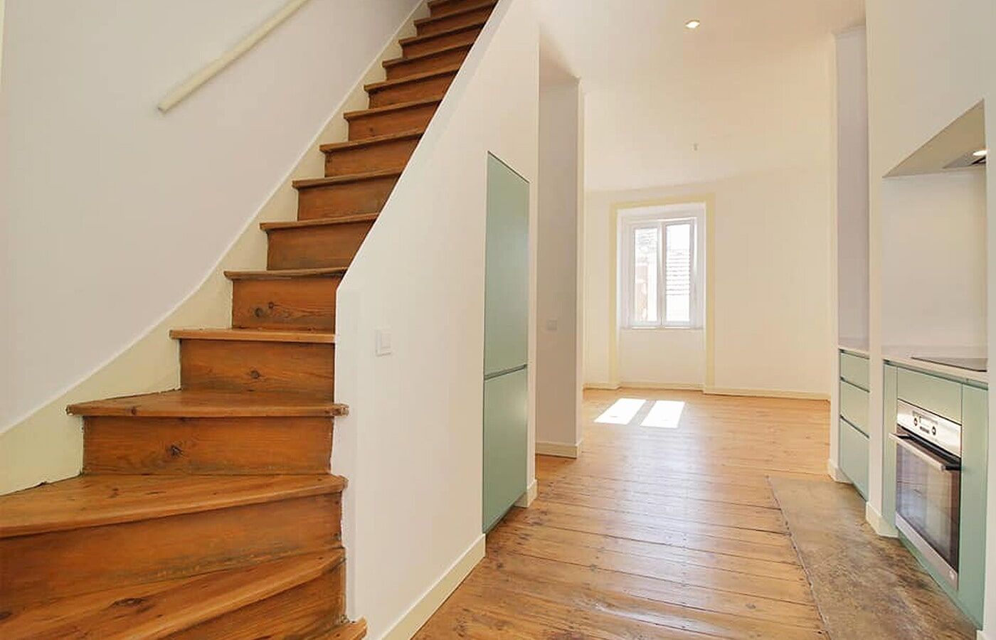 Escada de madeira em um corredor iluminado, com uma porta e uma vista parcial da cozinha.