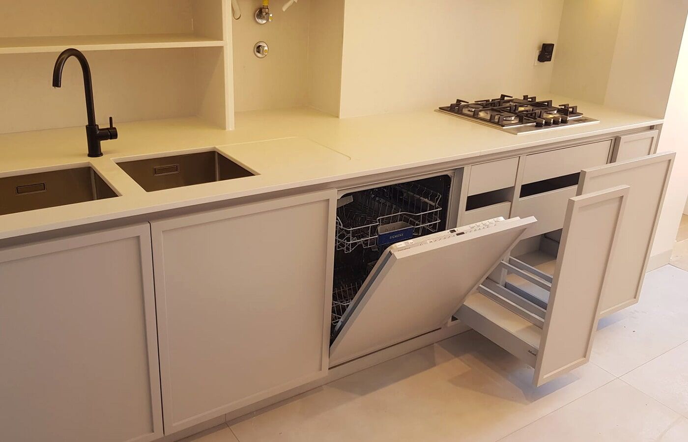 Cozinha branca com lava-louças aberta, torneira preta e pia de aço inoxidável.