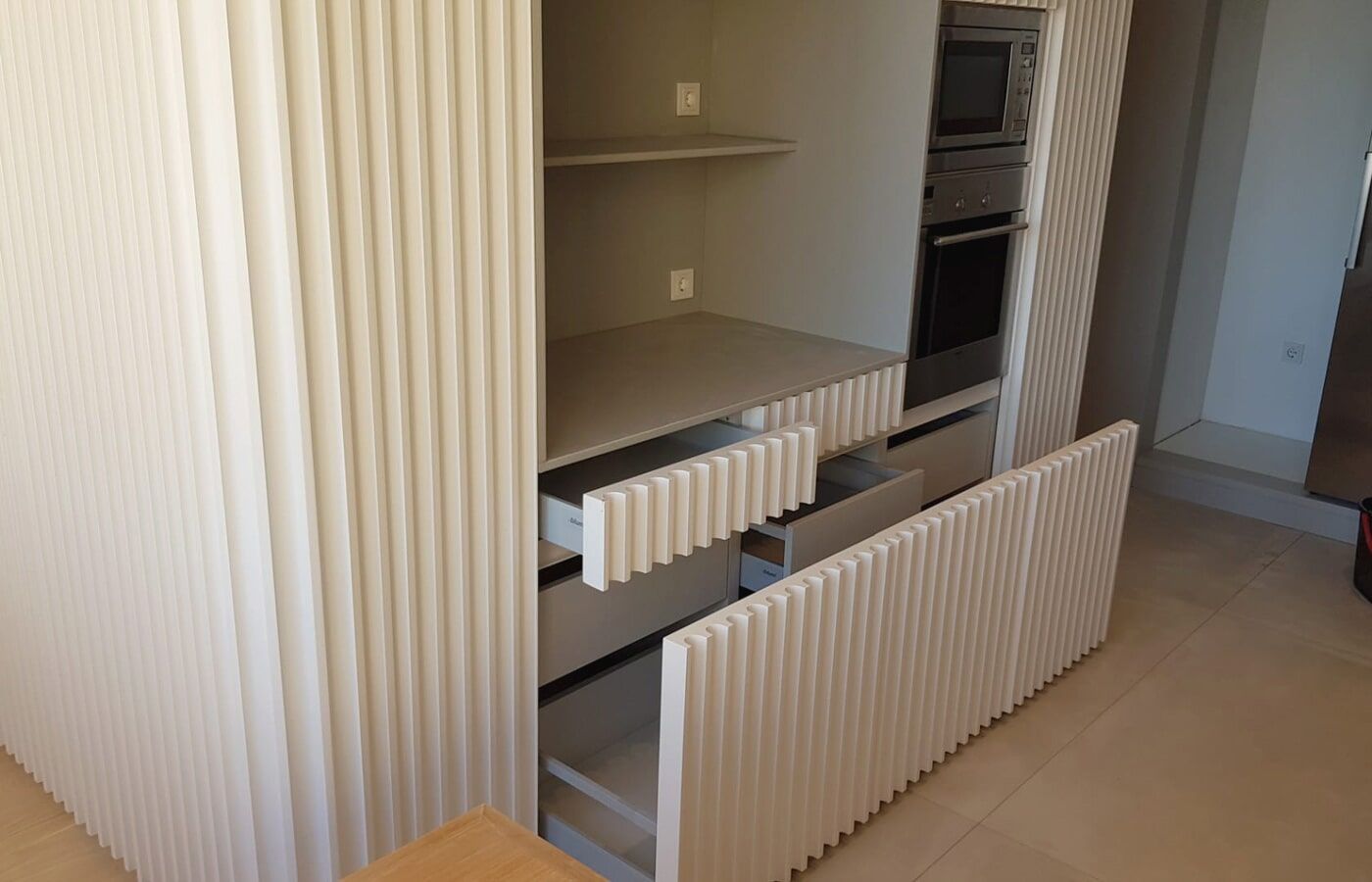 Armários de cozinha bege com linhas verticais texturizadas; gavetas e forno visíveis.