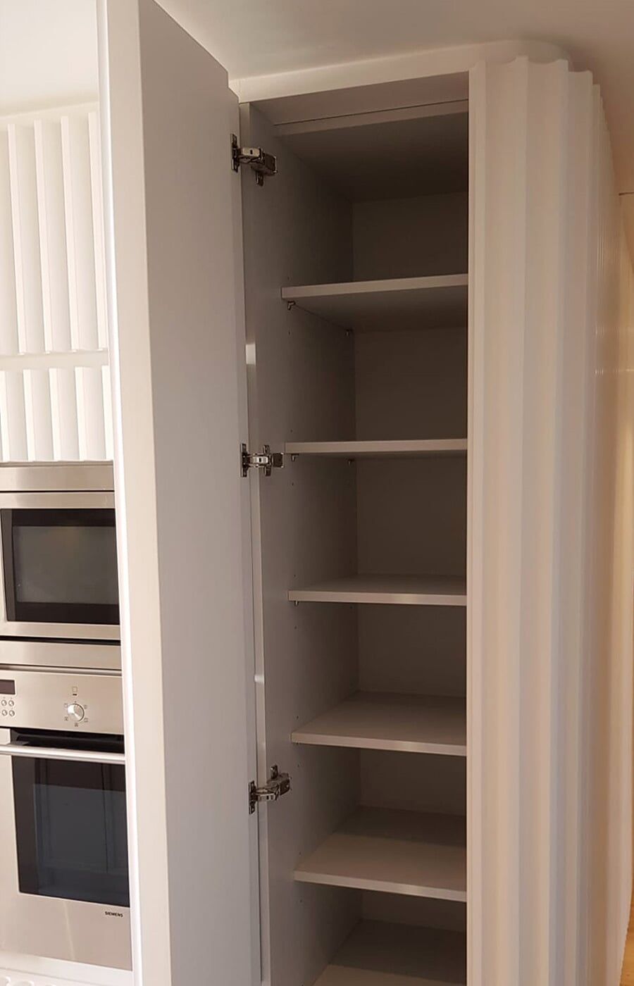 Armário de cozinha alto e cinza com porta aberta e várias prateleiras. Ao lado de uma parede branca texturizada e fornos.