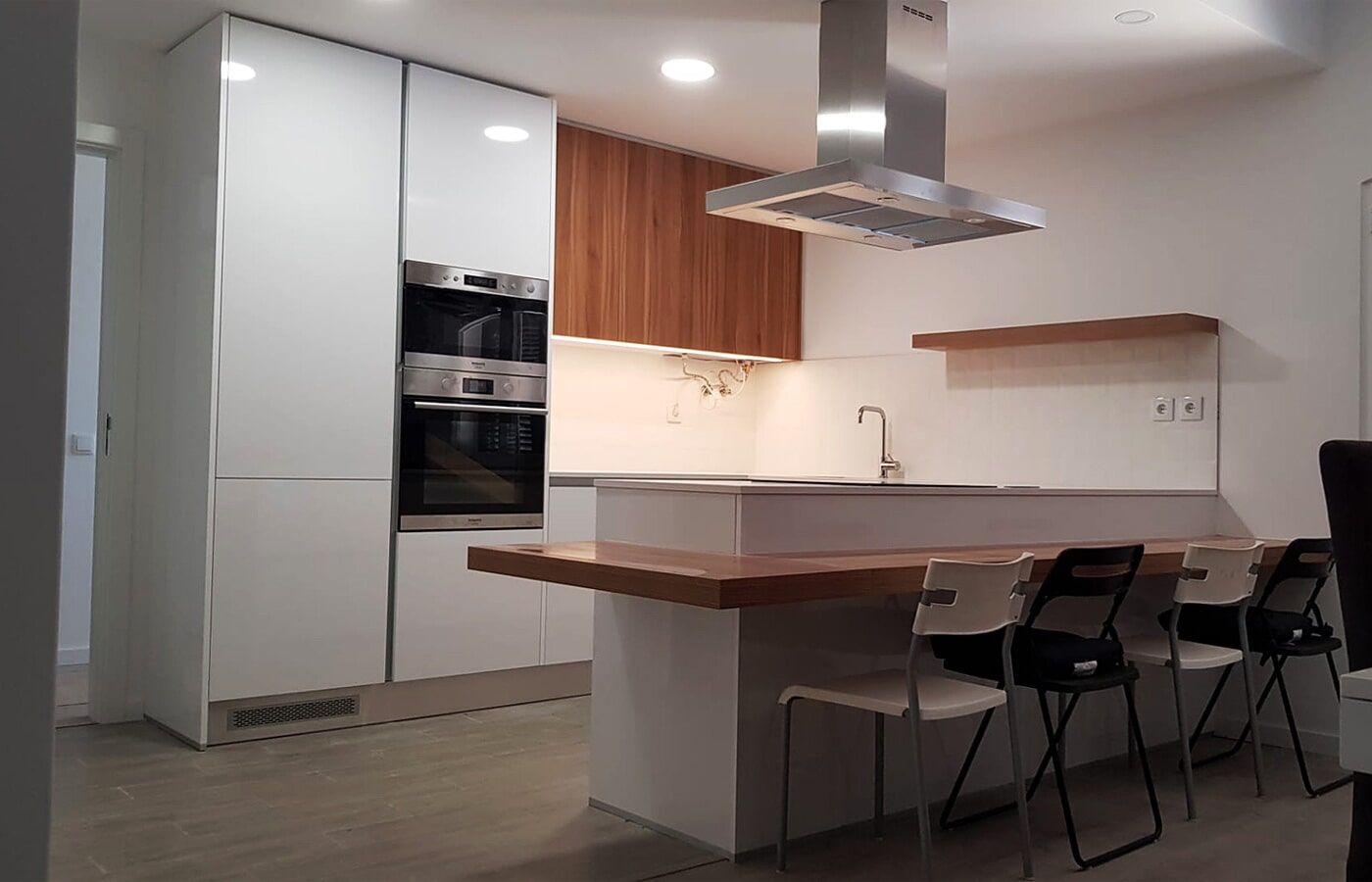 Cozinha moderna com armários brancos, detalhes em madeira e ilha com assentos. Coifa de aço inoxidável.