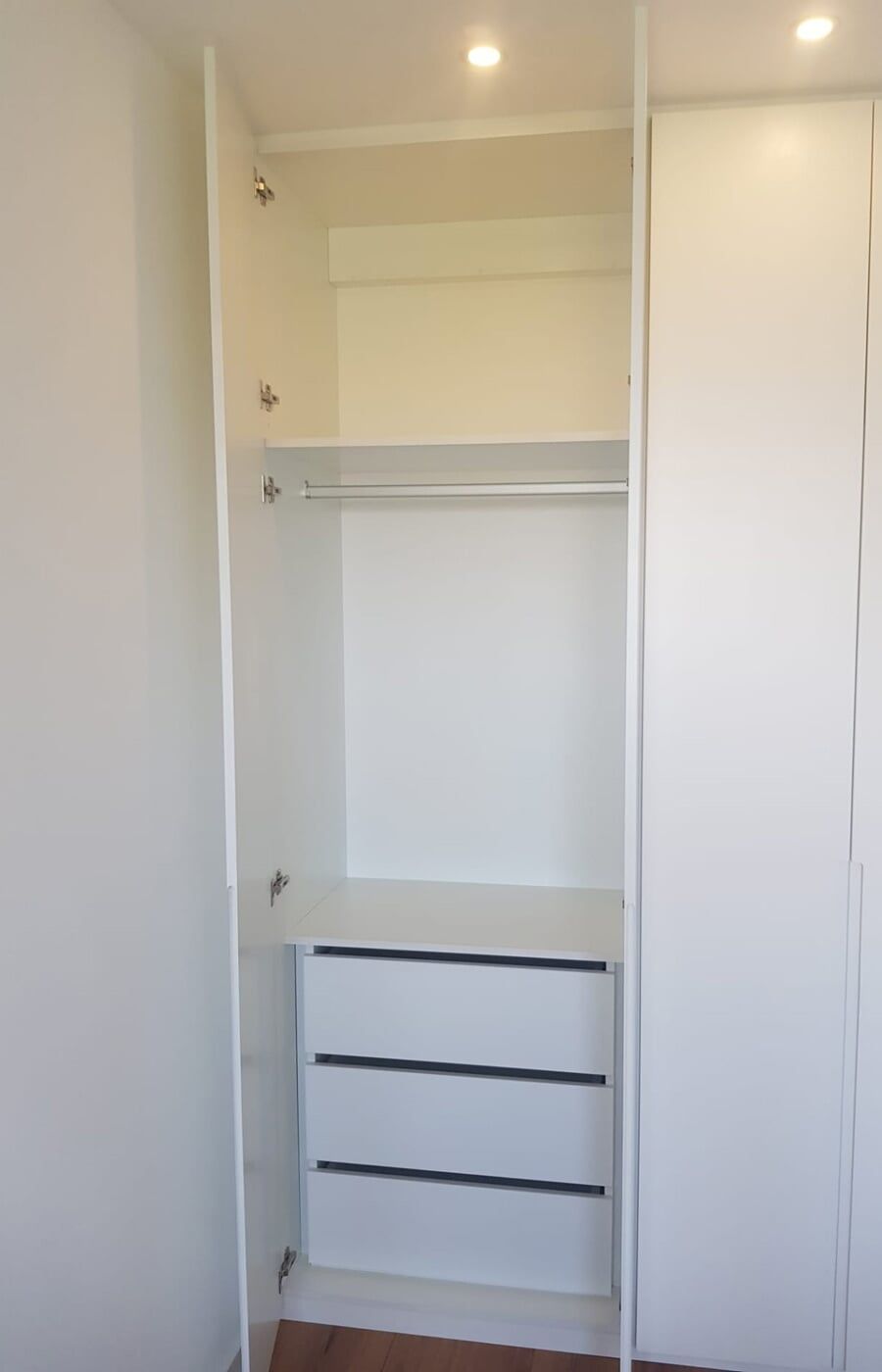 Interior de armário branco com porta aberta, apresentando um varão para pendurar roupas e três gavetas.