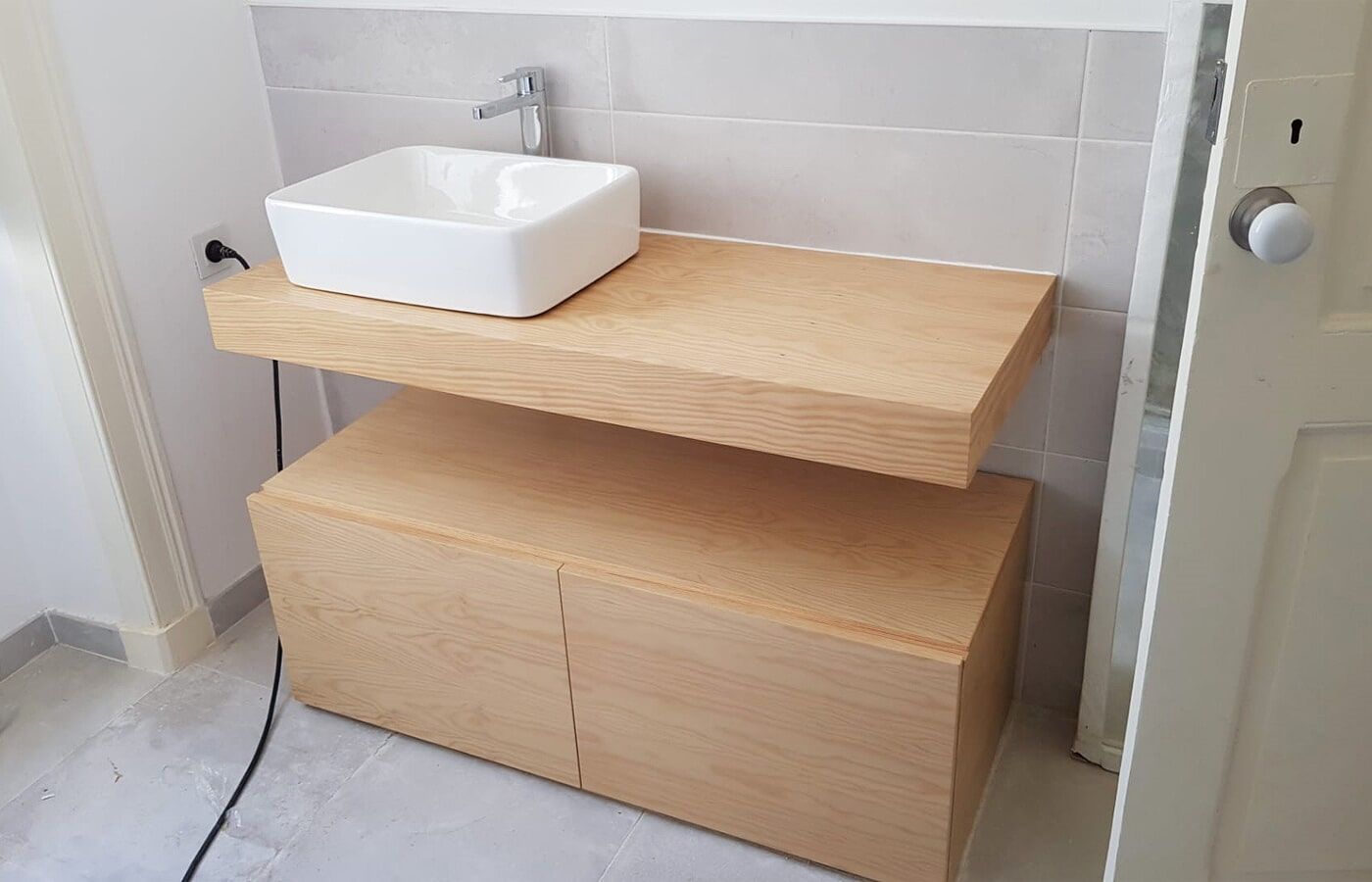 Móvel de banheiro em madeira com pia quadrada branca, armário de duas portas e parede revestida com azulejos cinza.