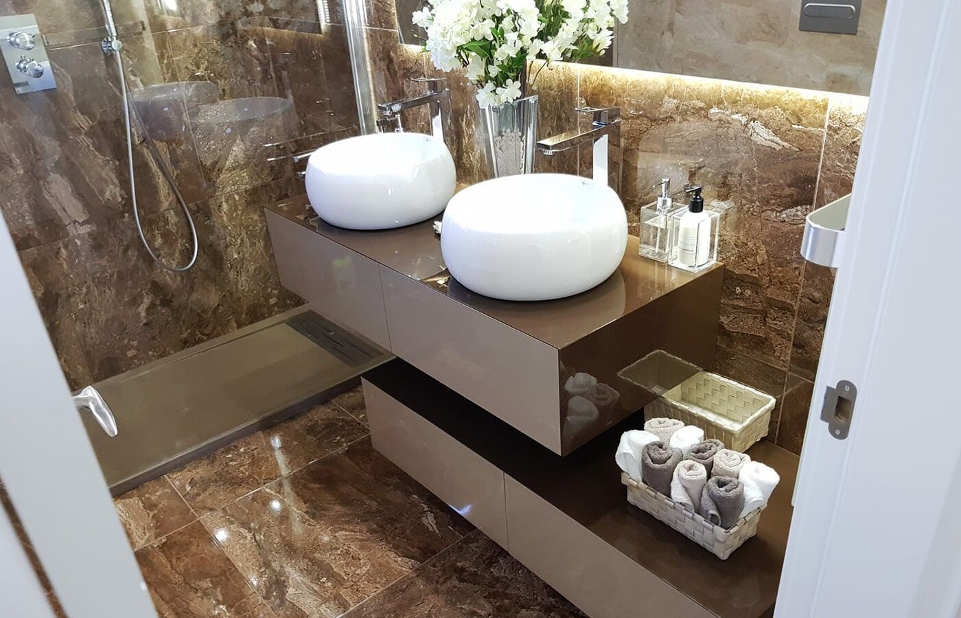 Banheiro moderno com paredes e piso de mármore marrom, bancada suspensa com duas cubas brancas e box de vidro.