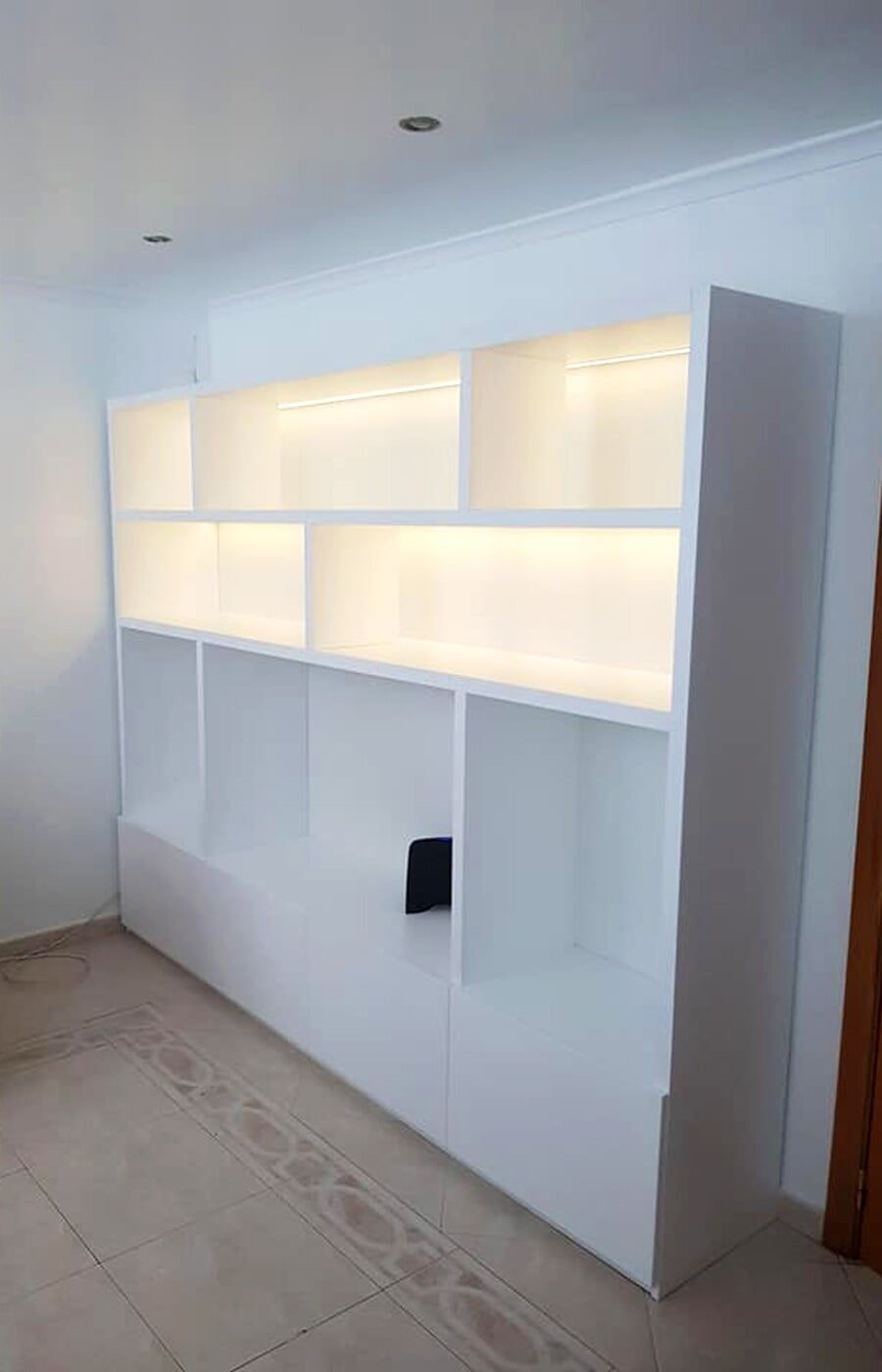 Estante embutida branca com iluminação integrada. A estante está instalada contra uma parede branca em um cômodo.